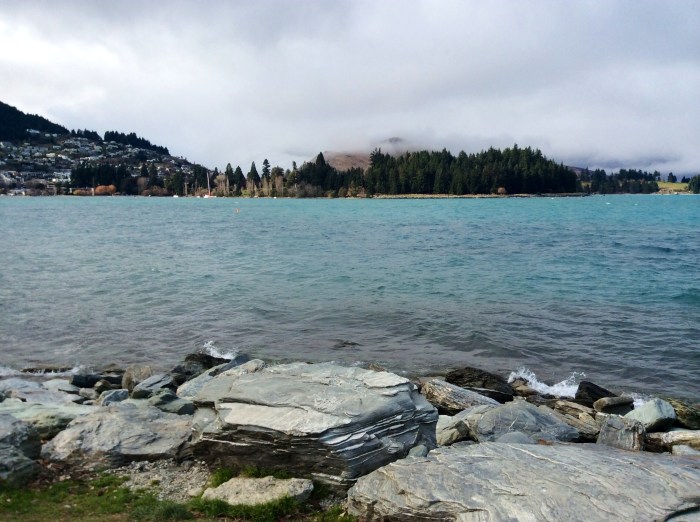 Les rives de la ville, formées par de petites plages de galets. Au loin, les jardins botanique de Queenstown et les fameux conifères